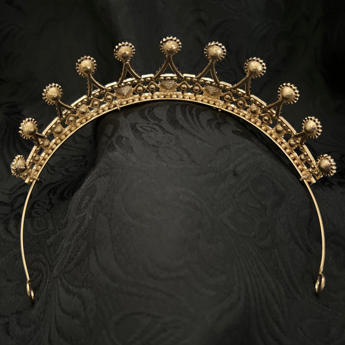 An ornate gold crown with decorative floral and geometric details, displayed on a textured black fabric background.
