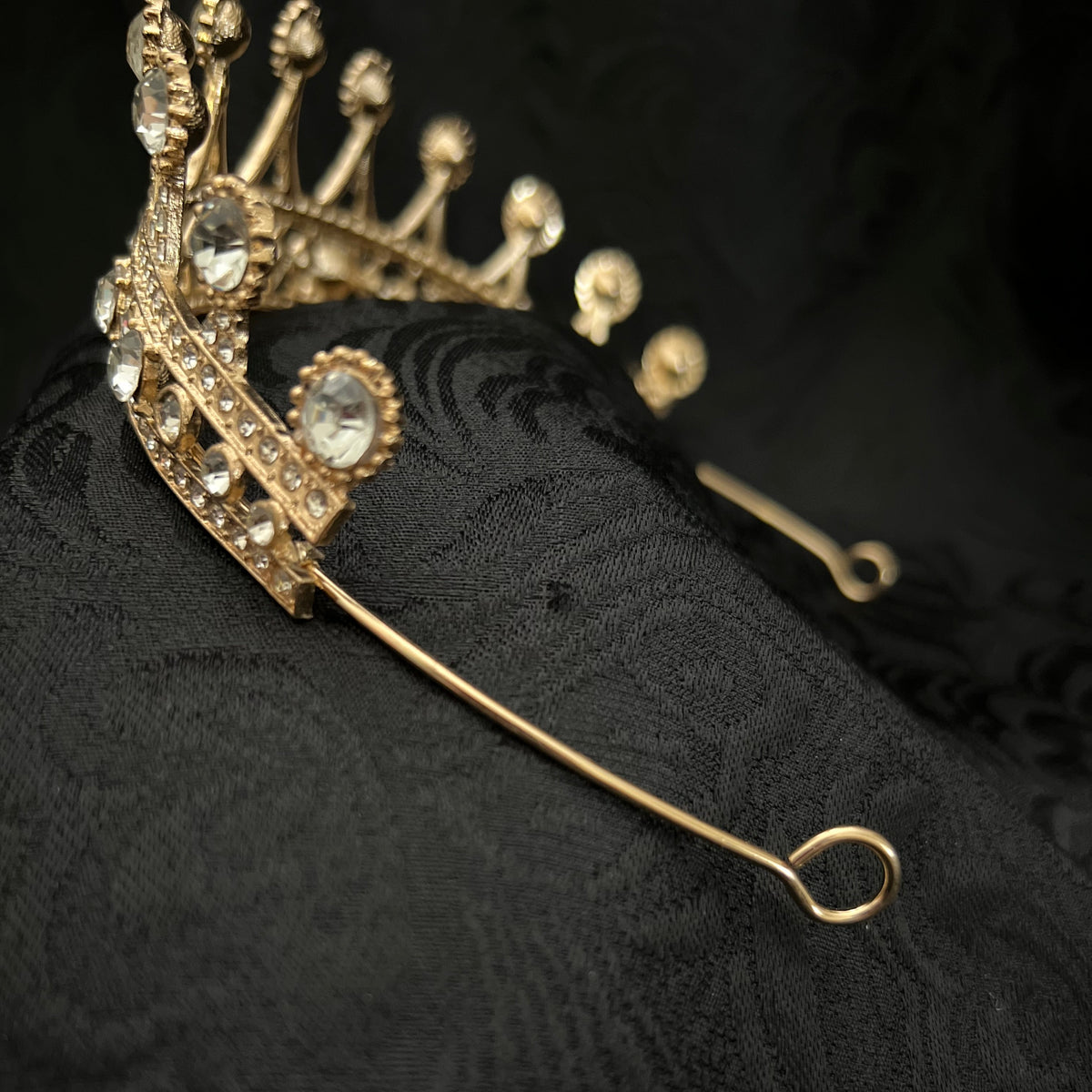 Gold crown hairpiece with clear rhinestones and a long looped tail, displayed on a textured black fabric background.