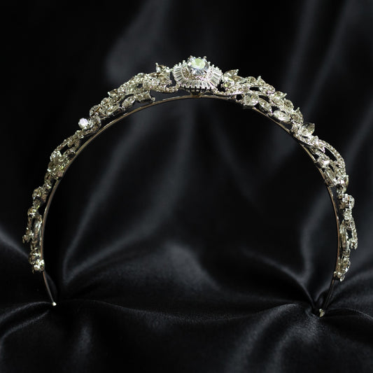 A silver-colored tiara with a central oval-shaped clear crystal surrounded by a halo of smaller baguette-style crystals, designed for hair adornment. bottom