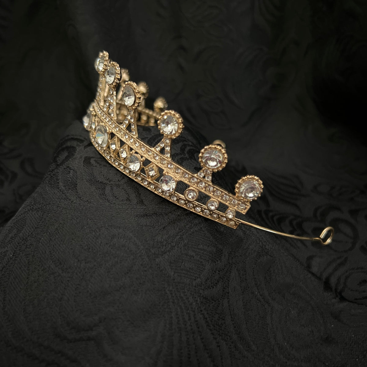 A gold crown tiara adorned with clear rhinestones, resting on a textured black fabric background.