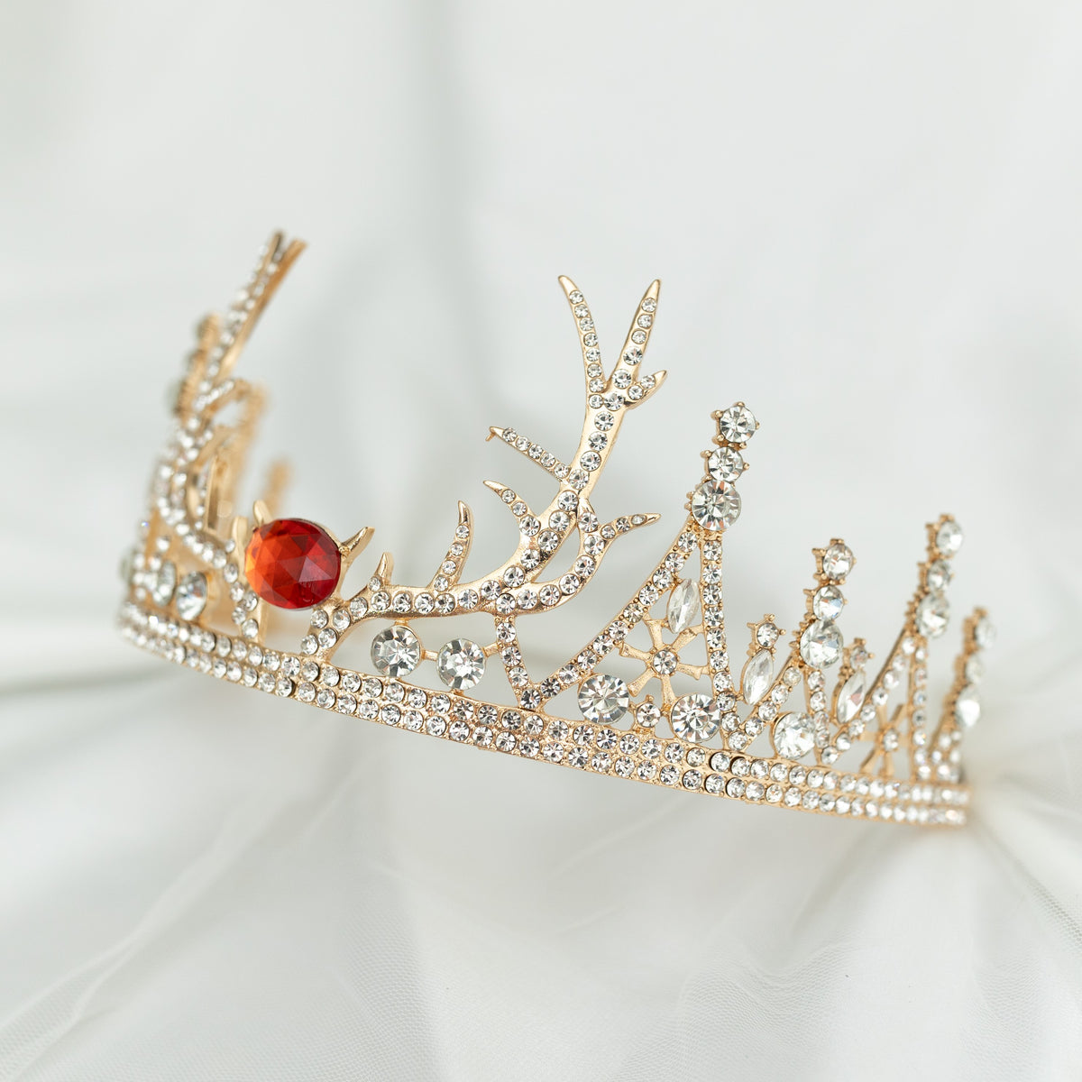 A sparkling tiara with a central red crystal, surrounded by clear crystals on a gold-colored base, designed to resemble antlers, set against a white background. Angled front view