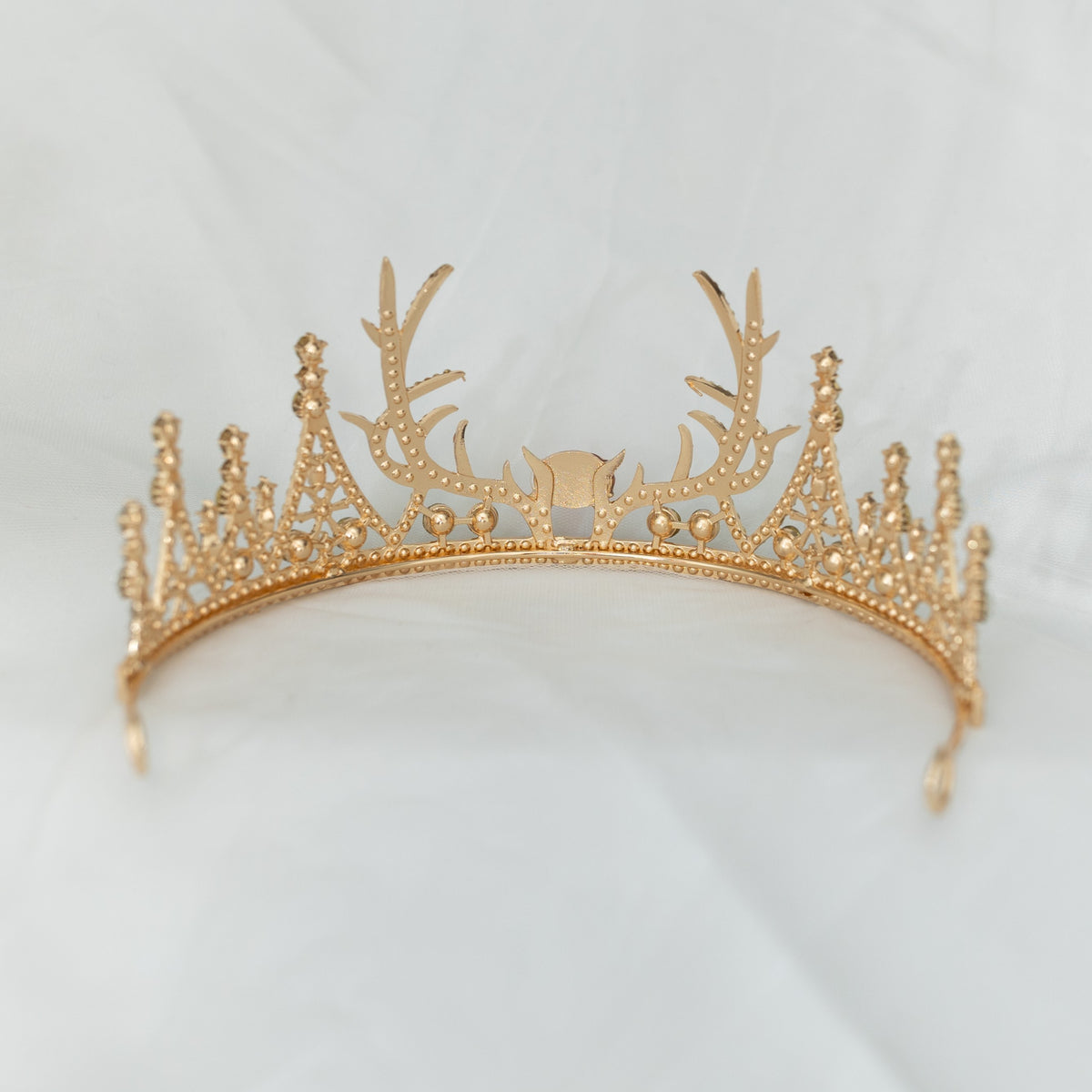 A sparkling tiara with a central red crystal, surrounded by clear crystals on a gold-colored base, designed to resemble antlers, set against a white background. back view