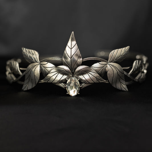 A silver tiara with a central clear stone, surrounded by laurel leaves, set against a black background. Front view