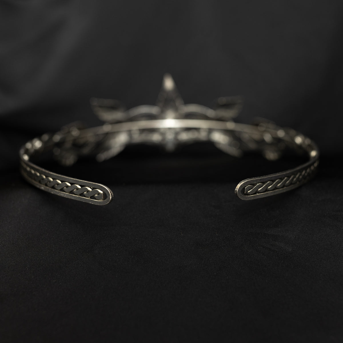 A silver tiara with a central clear stone, surrounded by laurel leaves, set against a black background. Rear view