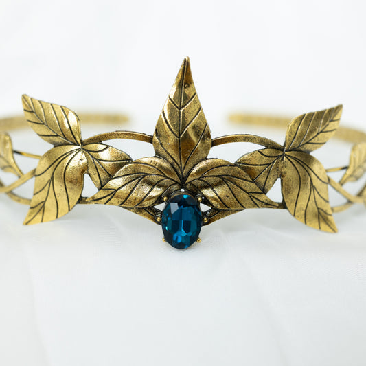 A golden tiara crown featuring a central dark blue stone, surrounded by laurel leaf detailing on a white background. Front close up