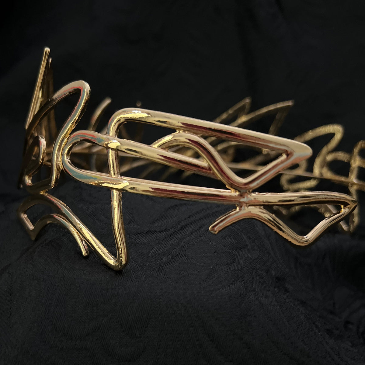 A gold abstract wire art piece with organic, flowing lines, displayed against a textured black fabric background.