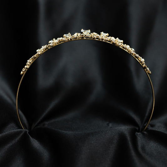 A delicate gold headband adorned with small clear rhinestones and a larger central crystal cluster, displayed against a draped black satin fabric background.