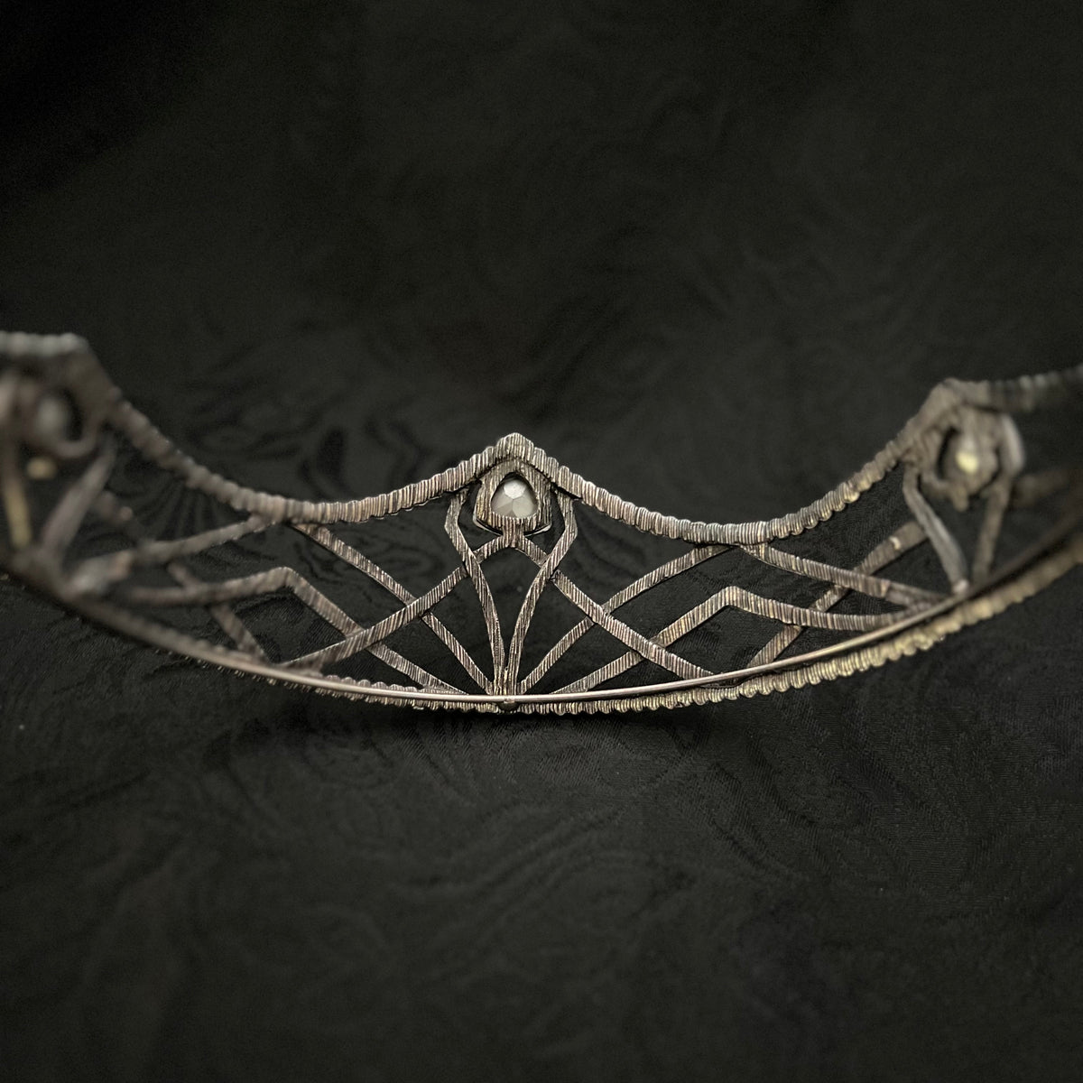 An ornate silver filigree tiara with geometric patterns and small pearl-like accents, displayed against a dark textured fabric background.