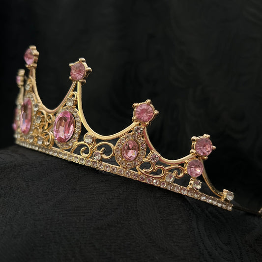 A gold tiara crown adorned with pink gemstones and small clear rhinestones, displayed on a textured black fabric background.r