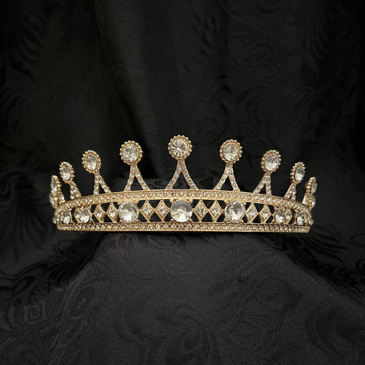 A gold crown adorned with clear gemstones, displayed on a textured black fabric background.