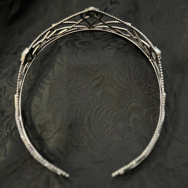 A silver-toned metal headband with delicate filigree detailing and small clear stones, displayed on a dark textured fabric background.