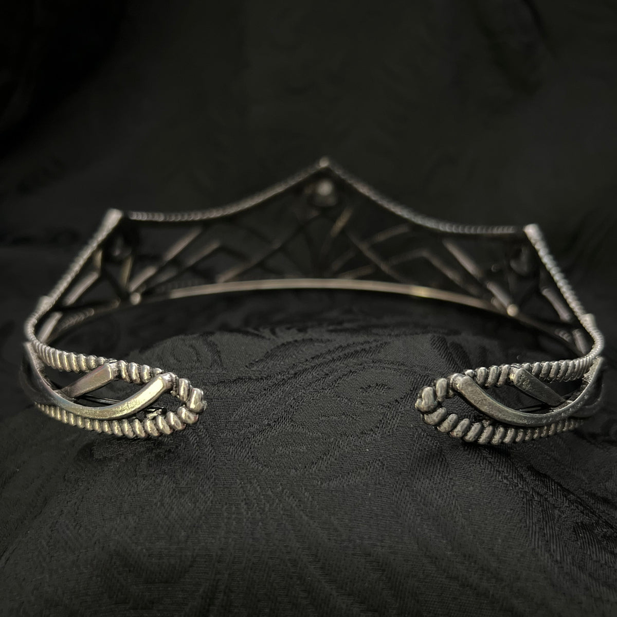 A close-up of a silver-toned metal garment accessory, likely a decorative brooch or part of a necklace, with textured scroll and geometric details, set against a black patterned fabric background.