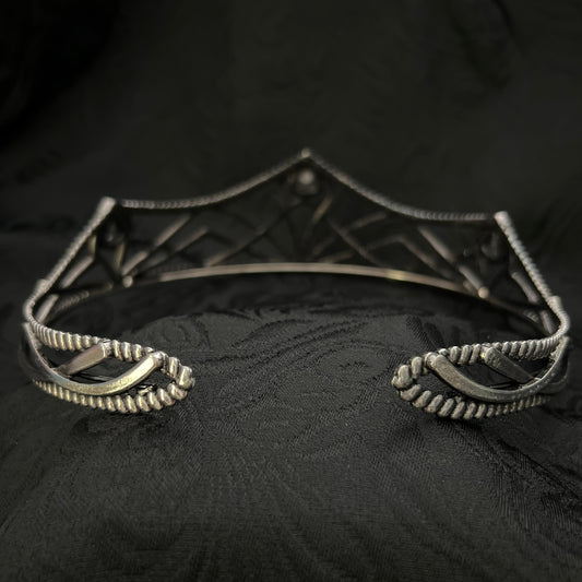 A close-up of a silver-toned metal garment accessory, likely a decorative brooch or part of a necklace, with textured scroll and geometric details, set against a black patterned fabric background.