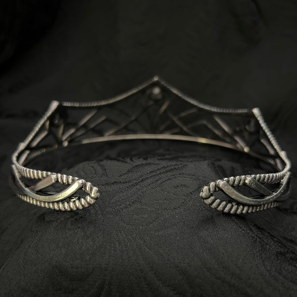 A close-up of a silver-toned metal garment accessory, likely a decorative brooch or part of a necklace, with textured scroll and geometric details, set against a black patterned fabric background.