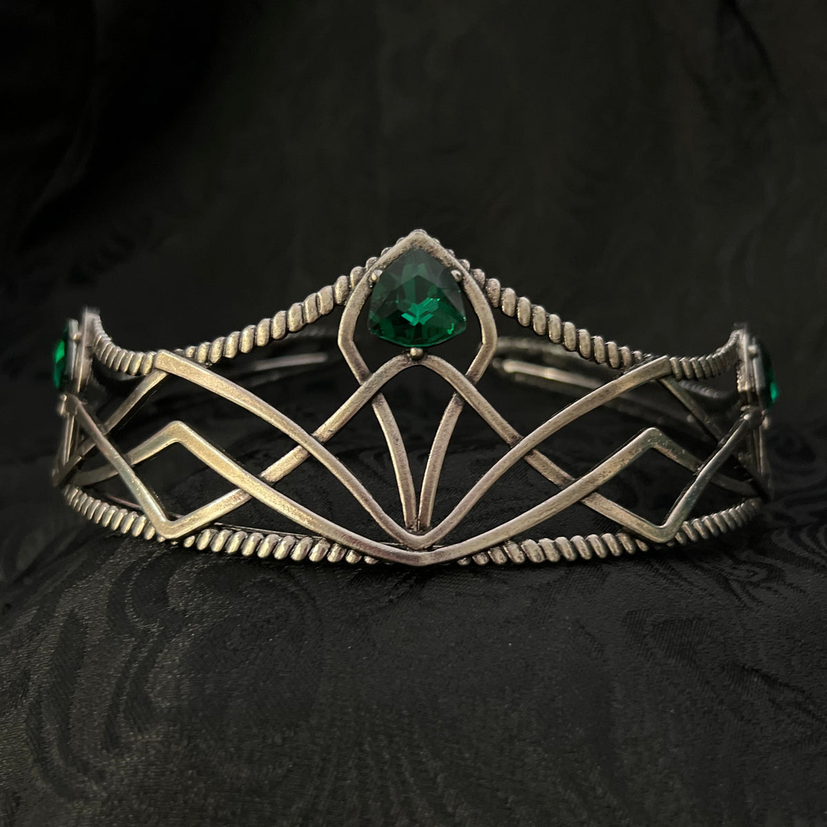 A silver-toned geometric cuff bracelet with a central green gemstone, displayed on a textured black fabric background.