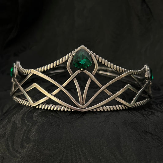 A silver-toned geometric cuff bracelet with a central green gemstone, displayed on a textured black fabric background.