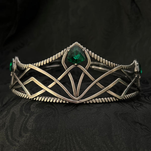 A silver-toned geometric cuff bracelet with a central green gemstone, displayed on a textured black fabric background.