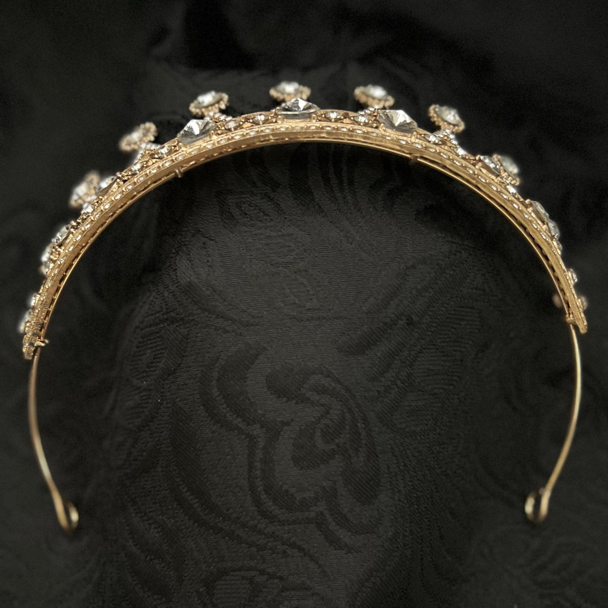 A gold tiara headband adorned with clear rhinestones, displayed on a textured black fabric background.