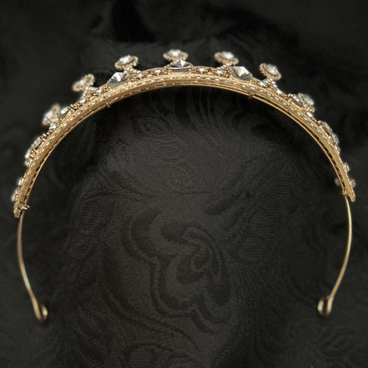 A gold tiara headband adorned with clear rhinestones, displayed on a textured black fabric background.
