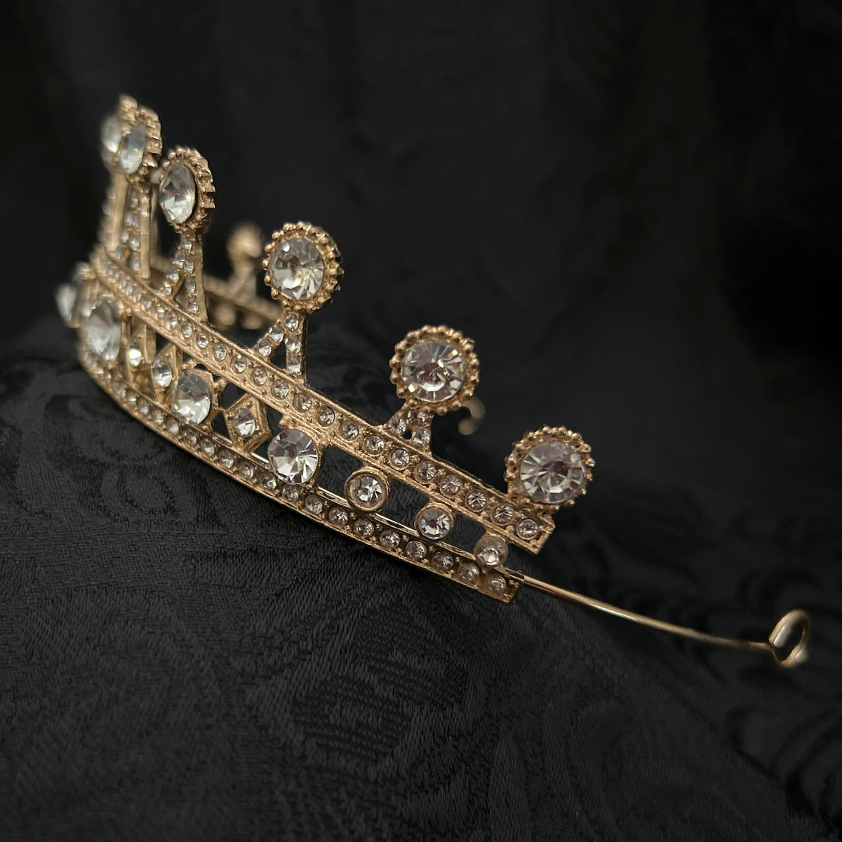 A gold crown tiara adorned with clear crystals, resting on a textured black fabric background.