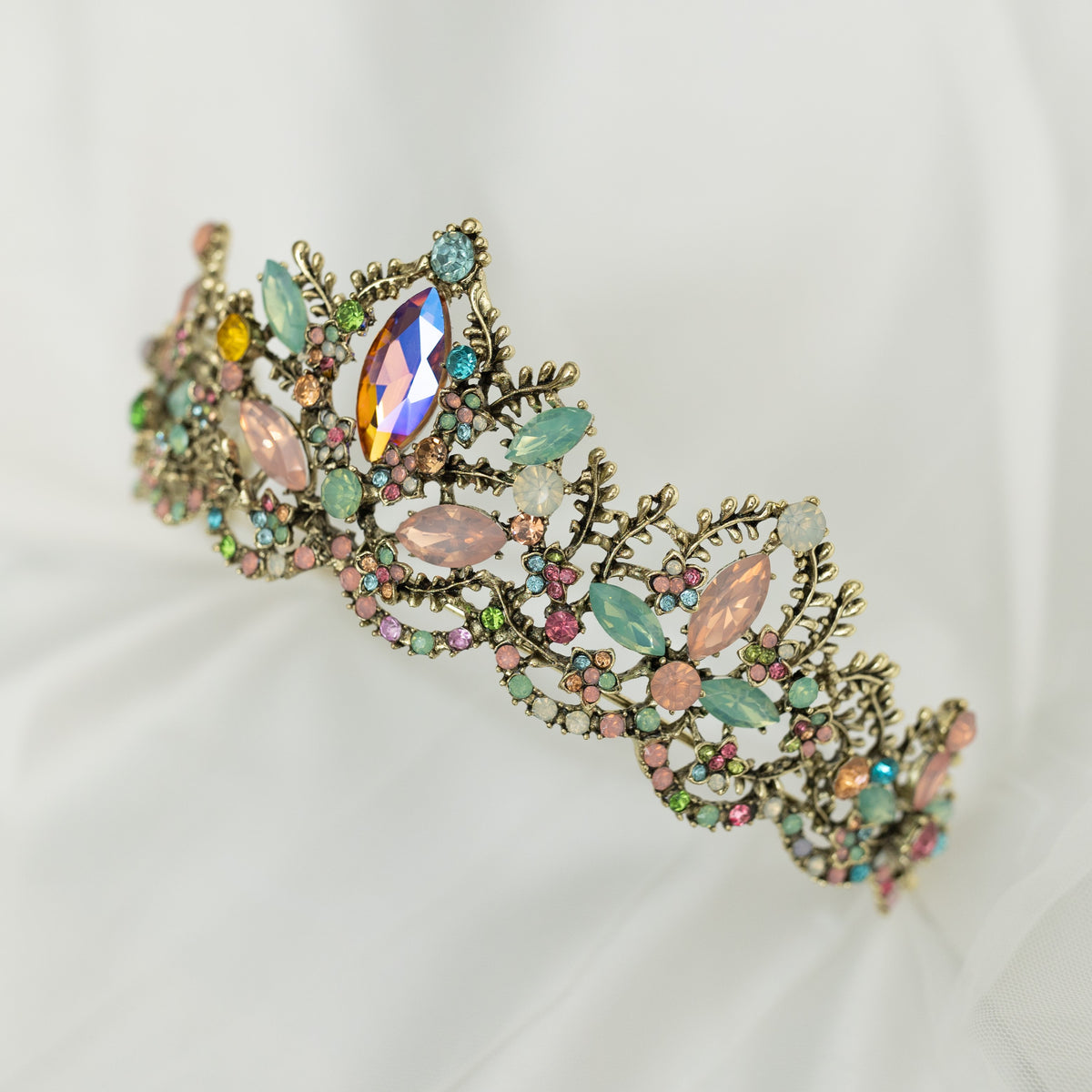 An antique gold-colored tiara adorned with large blue and pink crystals, and accented with pink, peach, blue, and turquoise color crystals. front angle view