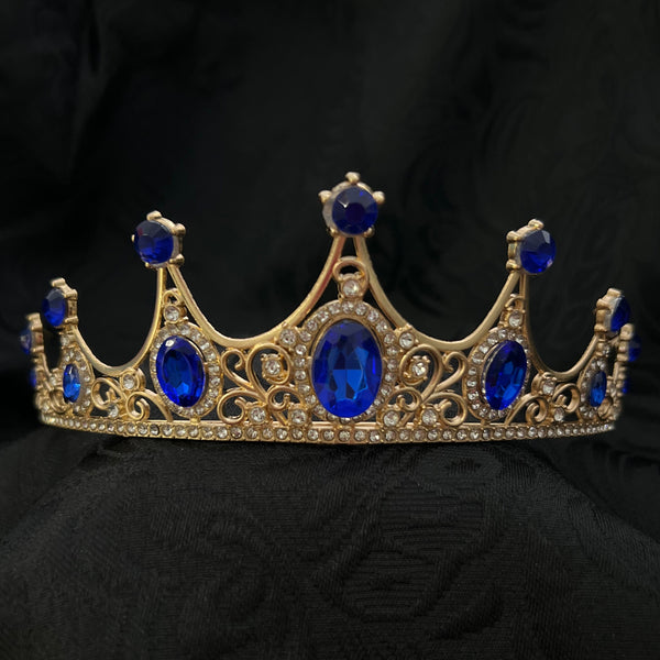 A gold crown tiara decorated with multiple oval and round deep blue faux jewels, displayed on a textured black fabric background.
