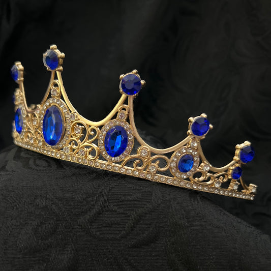 Gold crown brooch with blue gemstones, displayed on a black textured fabric background.