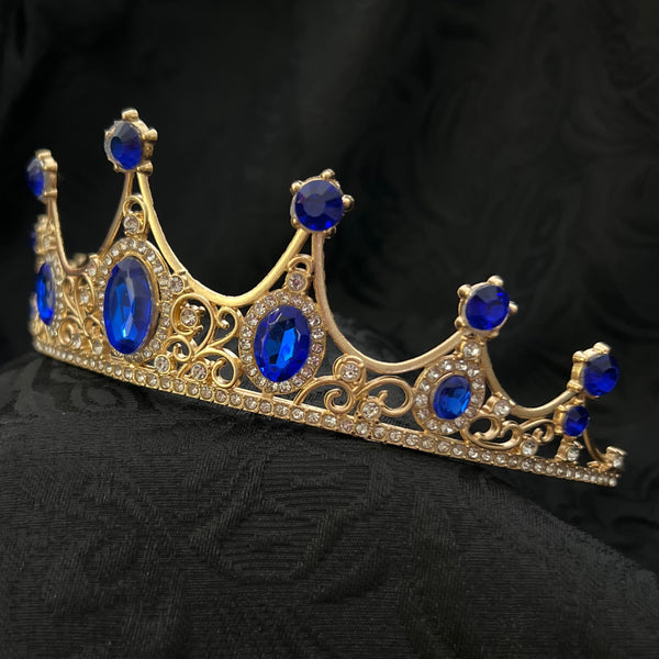 Gold crown brooch with blue gemstones, displayed on a black textured fabric background.