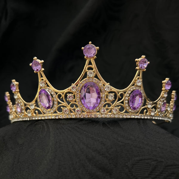 A gold crown-shaped jewelry piece with purple gemstones, photographed close-up against a black textured fabric background.
