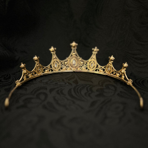 An ornate gold crown tiara with intricate filigree detailing and small central stones, displayed on a textured black fabric background.