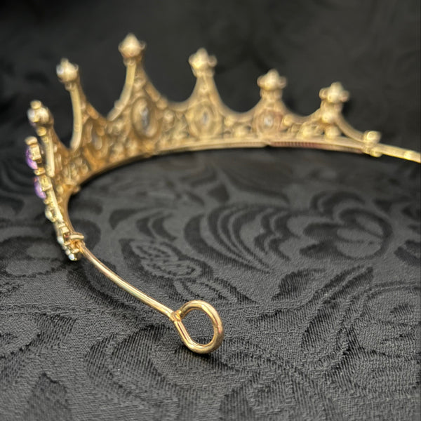 A gold crescent moon crown pendant with small clear rhinestones, shown close-up against a black damask fabric background.
