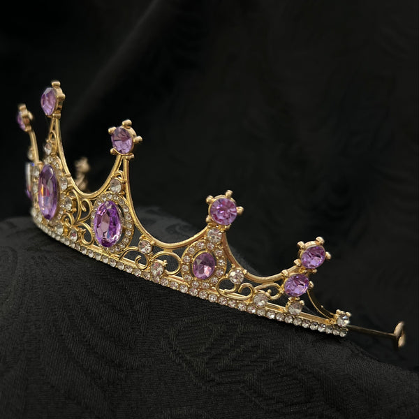 Gold crown tiara with purple gemstones and clear rhinestones, displayed on a black textured fabric background.