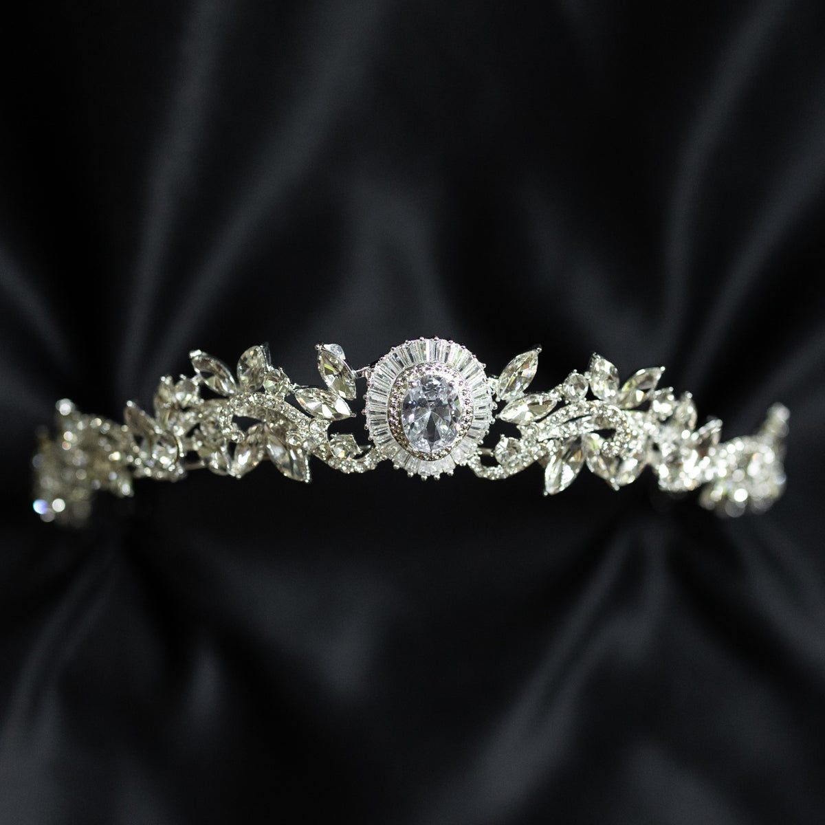 A silver-colored tiara with a central oval-shaped clear crystal surrounded by a halo of smaller baguette-style crystals, designed for hair adornment.