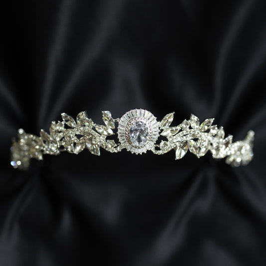 A silver-colored tiara with a central oval-shaped clear crystal surrounded by a halo of smaller baguette-style crystals, designed for hair adornment.