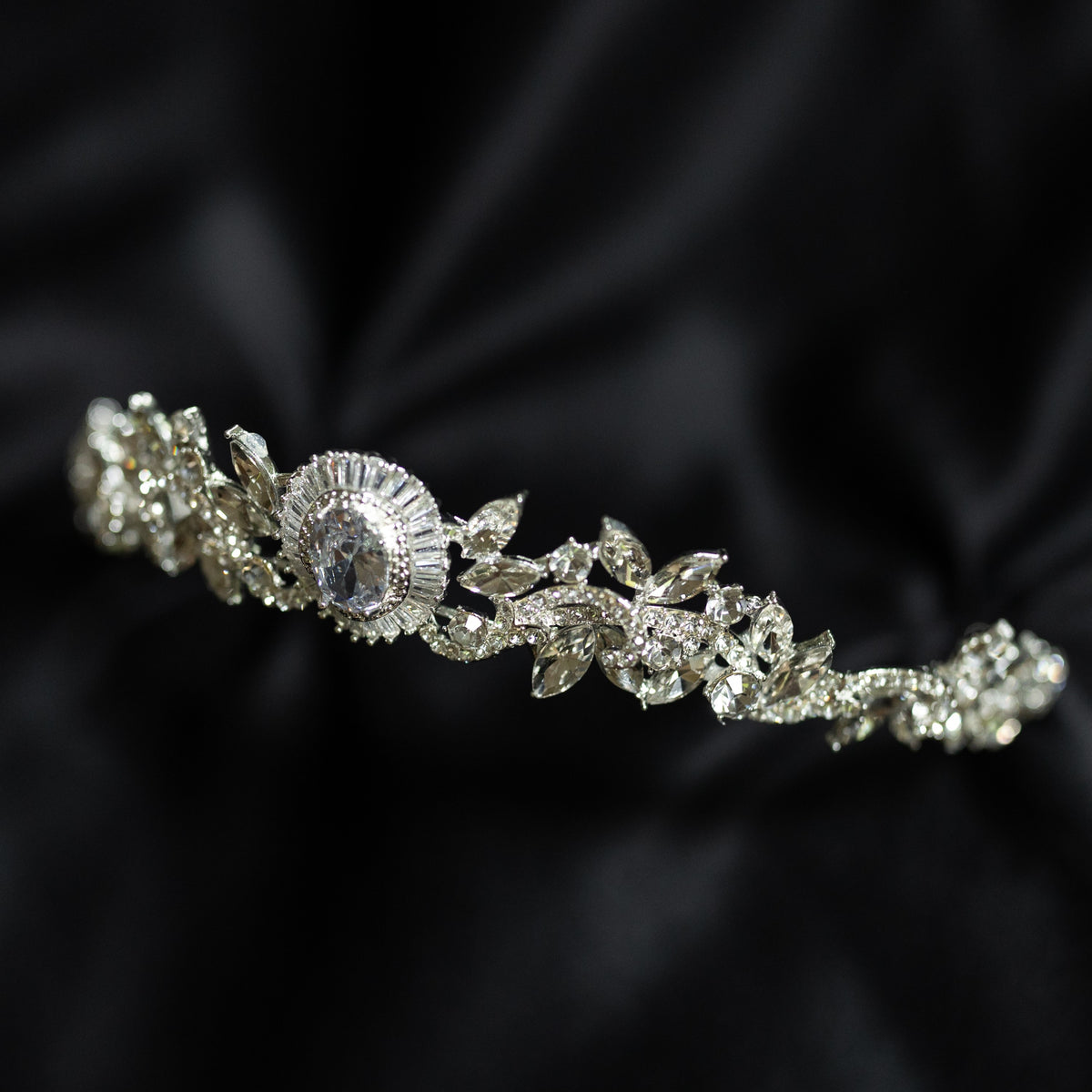 A silver-colored tiara with a central oval-shaped clear crystal surrounded by a halo of smaller baguette-style crystals, designed for hair adornment. front angle