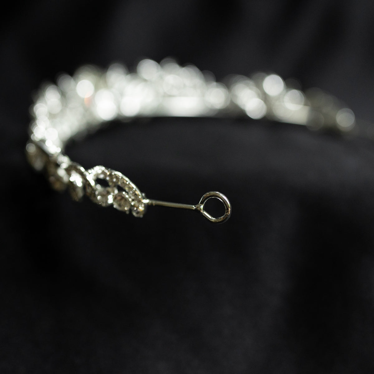 A silver-colored tiara with a central oval-shaped clear crystal surrounded by a halo of smaller baguette-style crystals, designed for hair adornment. looped end
