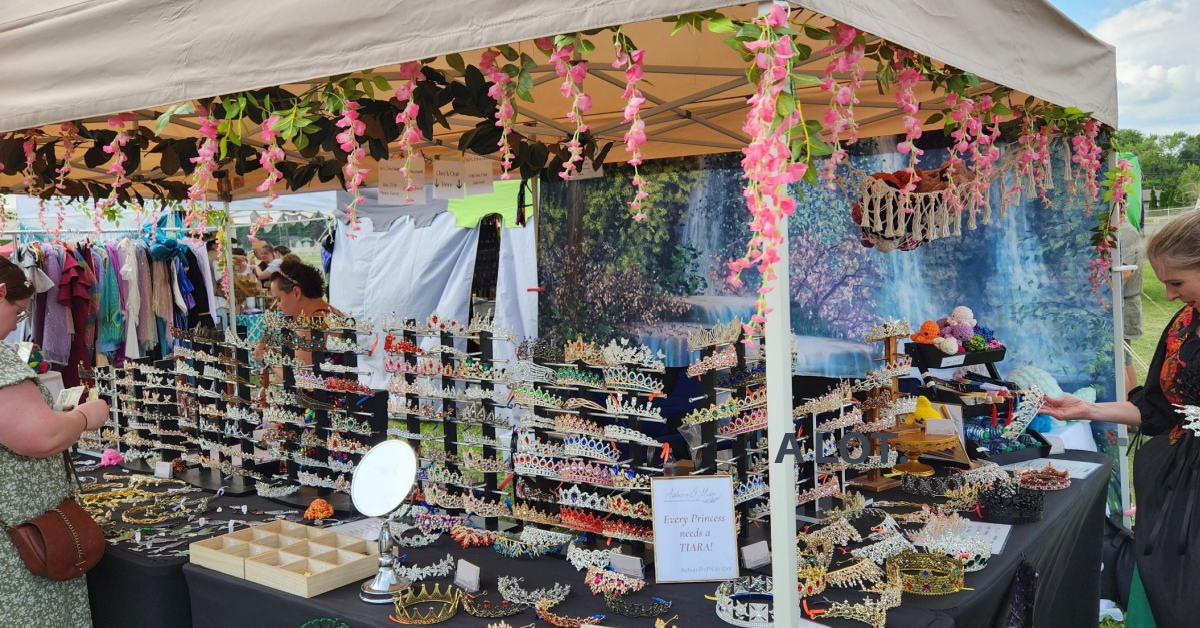 Stall with various items for sale under a canopy with floral decorations - Auburn And Main’s festival in person show tent with tiaras displayed for sale 