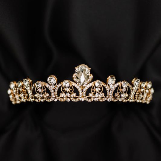 A gold-colored metal tiara with clear crystal embellishments, displayed against a black background.