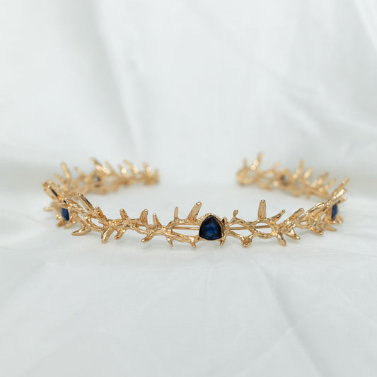 A gold colored metal crown with spikes and blue faux sapphire stones form a disconnected circle, displayed against a white background.