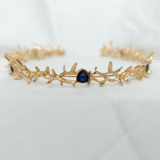 A gold colored metal crown with spikes and blue faux sapphire stones form a disconnected circle, displayed against a white background. close up