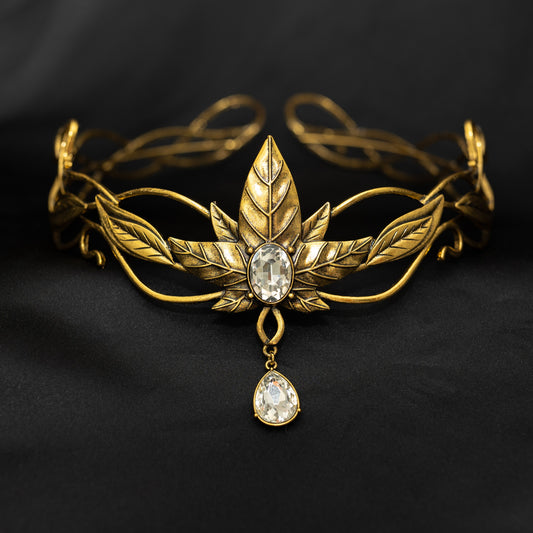 A golden tiara featuring a central design with five laurel leaves and a large clear crystal, with additional leaf details and a dangling crystal.