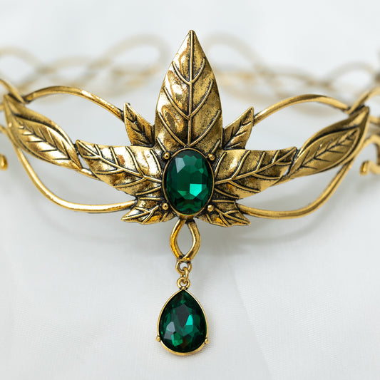 A golden tiara featuring a central green crystal with additional leaf detailing and a dangling green crystal, designed for adult cosplay or as a decorative piece. Front close up view 