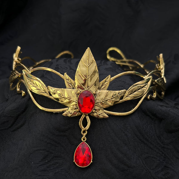 An ornate gold-toned metal brooch or pendant featuring engraved leaf motifs and two red gemstones, one round and one teardrop, displayed on a textured black fabric background.