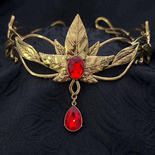 An ornate gold-toned metal headband featuring leaf and vine motifs, decorated with two red teardrop gemstones, displayed on a textured black fabric background.