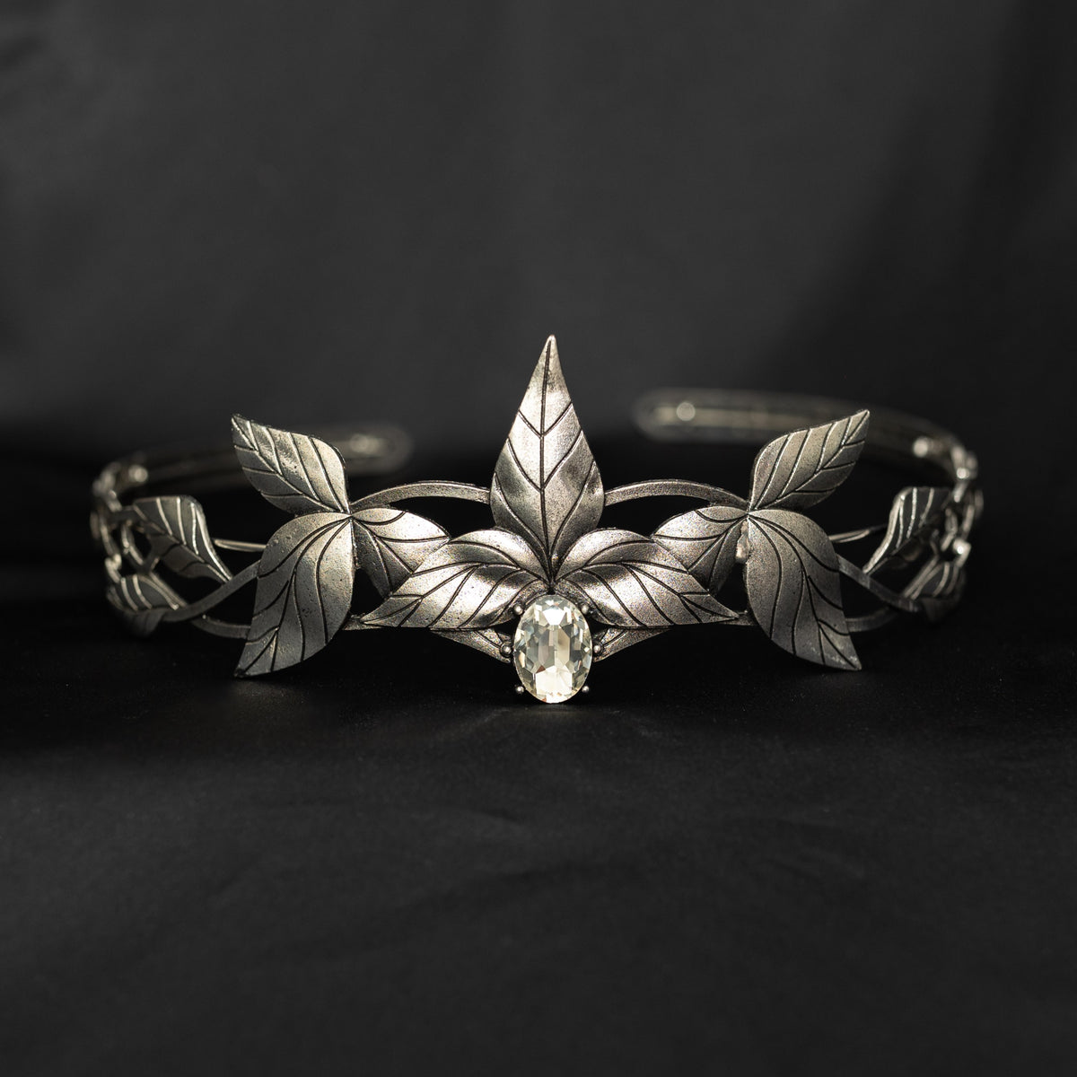 A silver tiara with a central clear stone, surrounded by laurel leaves, set against a black background. Front view 