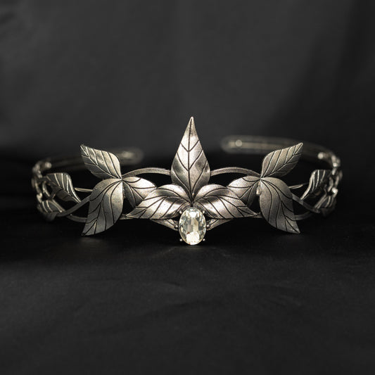 A silver tiara with a central clear stone, surrounded by laurel leaves, set against a black background. Front view 