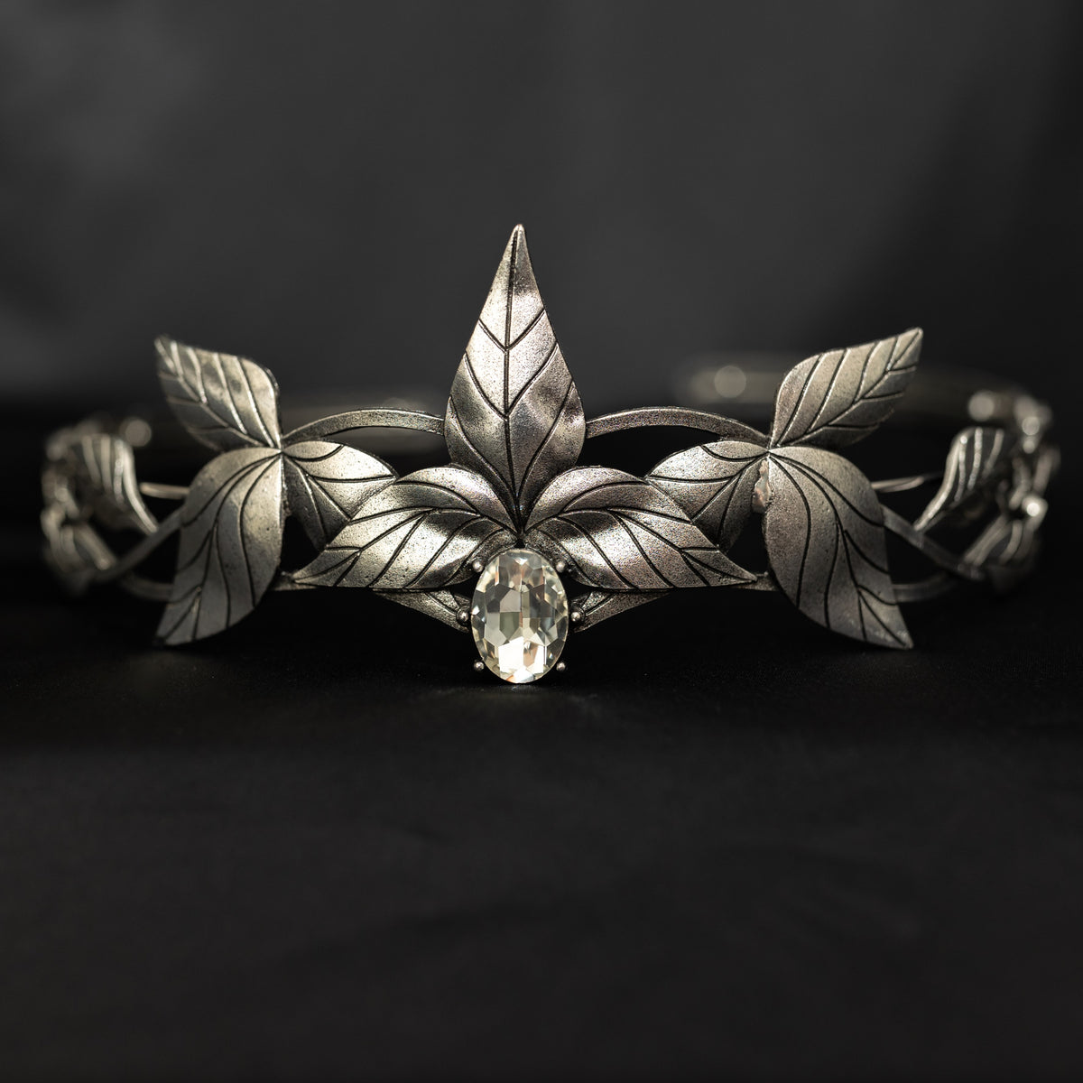 A silver tiara with a central clear stone, surrounded by laurel leaves, set against a black background. Front view 