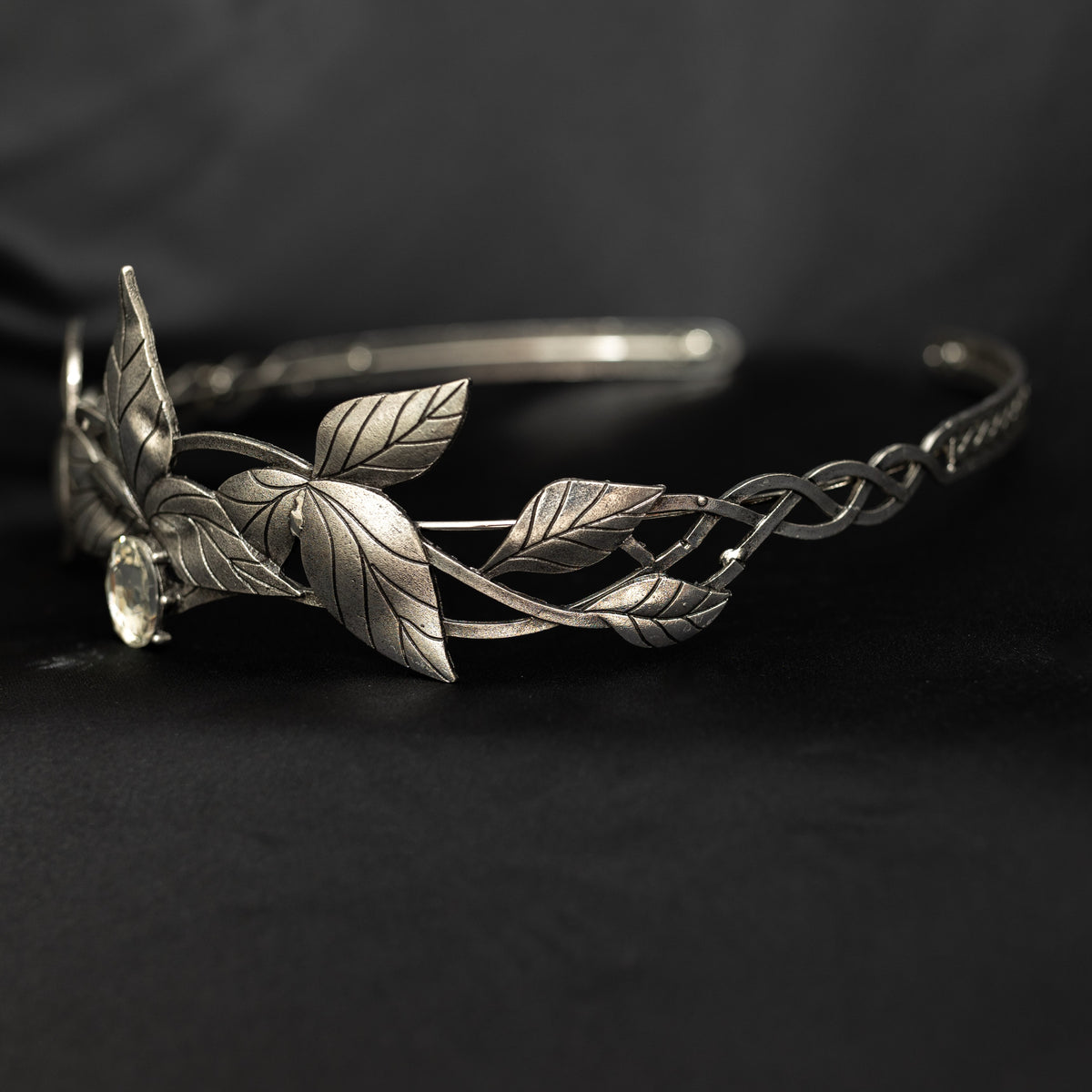 A silver tiara with a central clear stone, surrounded by laurel leaves, set against a black background. Front side view 