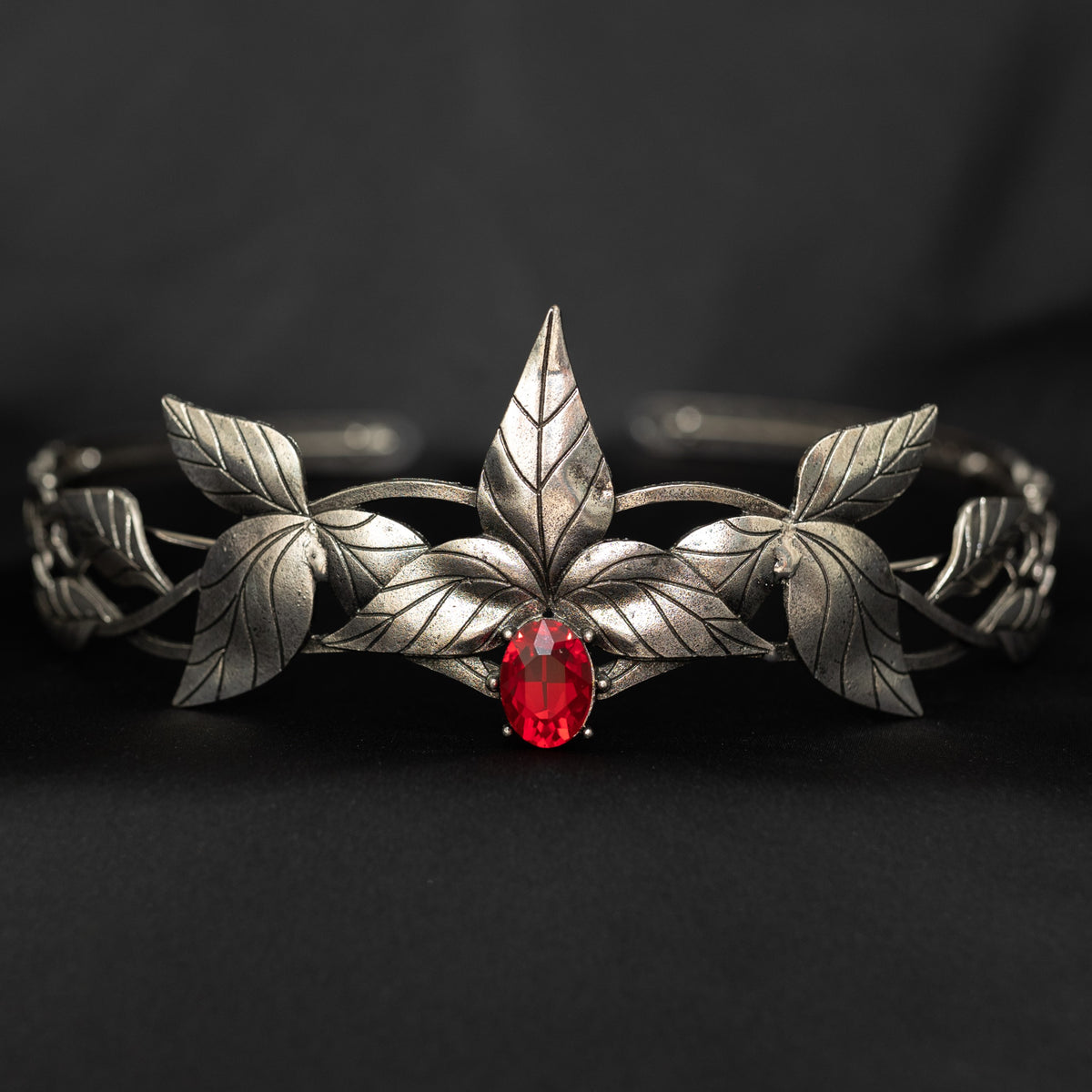 A silver-colored tiara with laurel leaf patterns and a central red stone, presented against a black background. Front view 