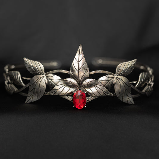 A silver-colored tiara with laurel leaf patterns and a central red stone, presented against a black background. Front view 